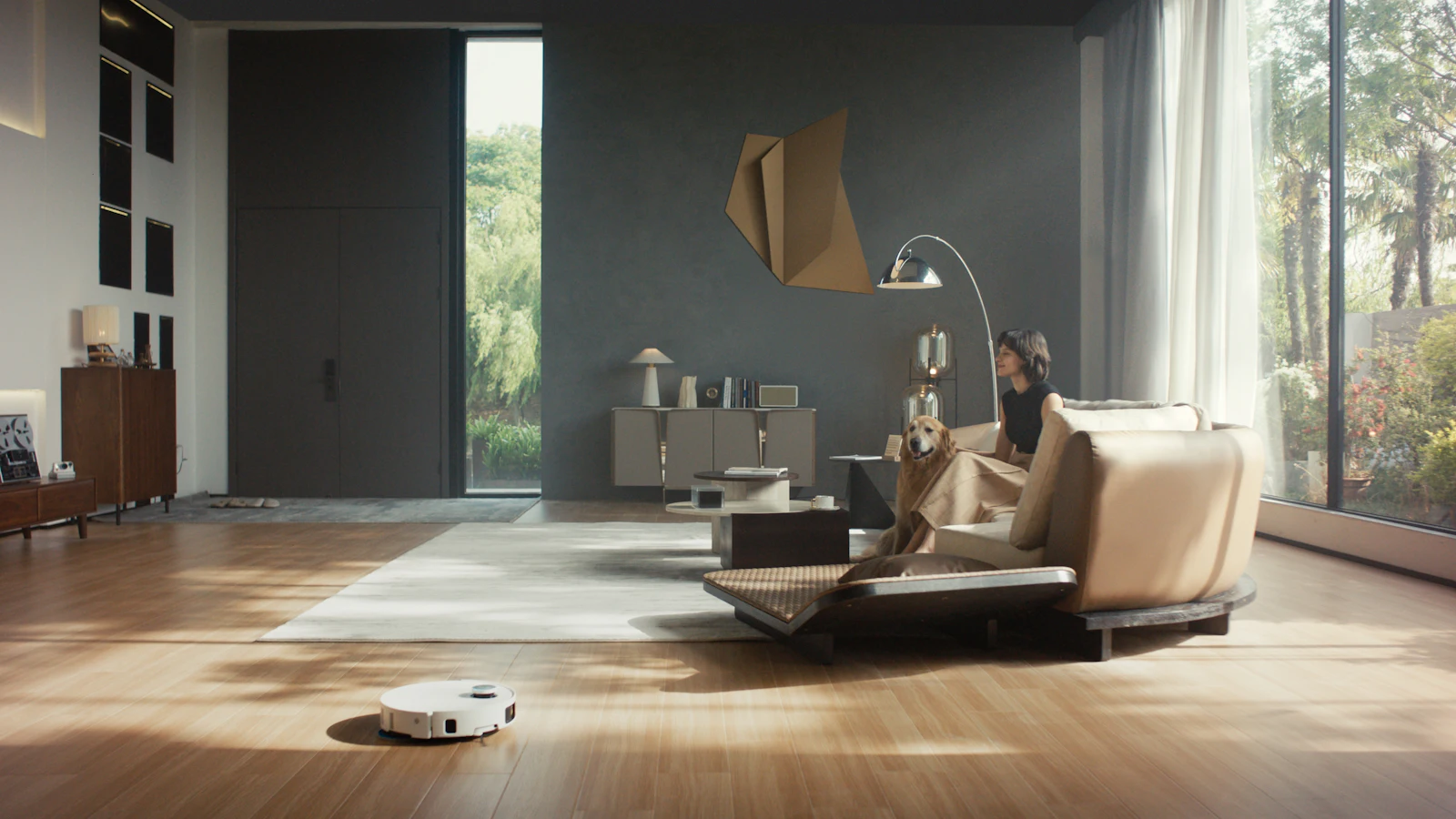 Un robot aspirateur autonome nettoie le parquet d'un salon lumineux pendant qu'une femme et son chien se détendent sur le canapé